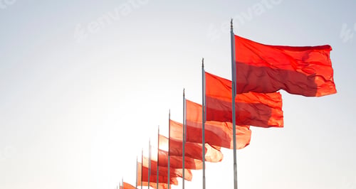 Preview: Row of bright red flags on flagpoles fluttering in the wind, clear sky.