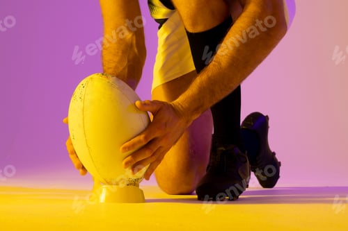 Preview: Caucasian male rugby player crouching with rugby ball over pink lighting