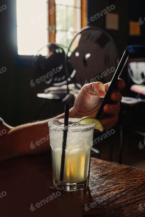 Preview: Light from a window on a margarita in a dark restaurant and a male hand holding a mobile phone