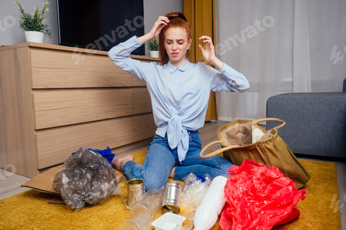 Preview: unhappy redhaired ginger girl sorting waste:bottle,paper and plastic sitting at floor carpet