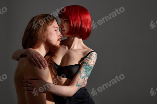 Preview: A young attractive woman with bright ruby hair and a tattoo in black lingerie embraces a man with