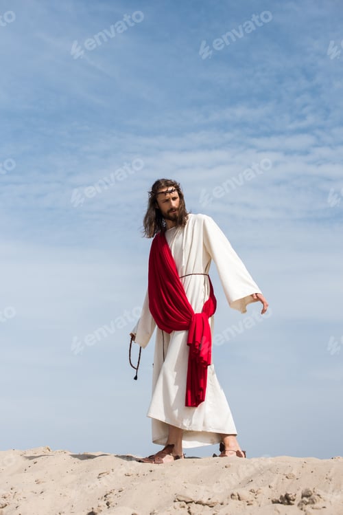 Preview: Jesus in robe, red sash and crown of thorns holding rosary and walking on sandy hill in desert