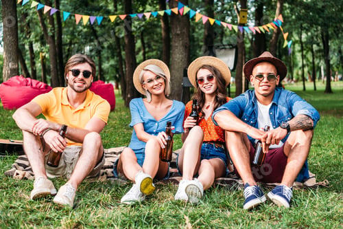 Preview: happy multiracial friends with beer resting on blanket in summer park