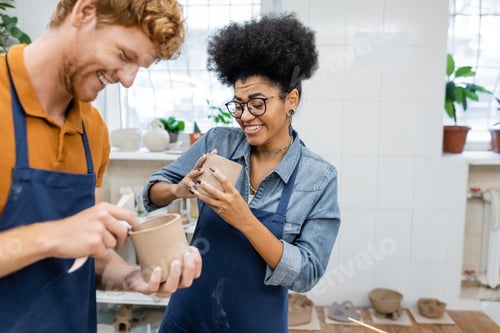 Preview: cheerful african american woman in eyeglasses molding clay cup with happy redhead boyfriend in