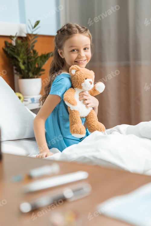 Preview: Little patient with teddy bear sitting on bed in hospital