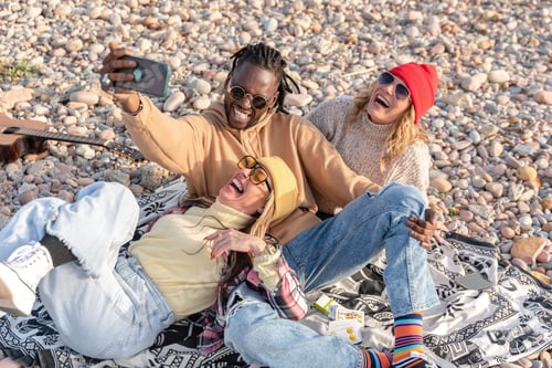 Preview: Group of happy multi-ethnic friends taking a selfie while having fun on a pebble beach
