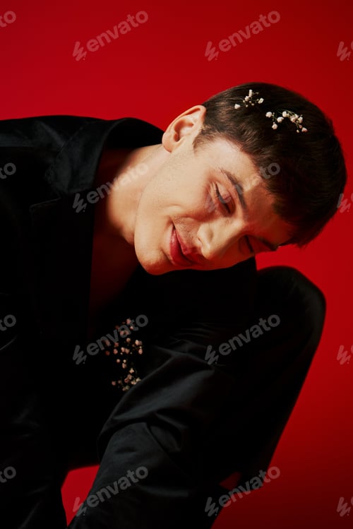 Preview: cheerful young man in black velvet jacket with flowers in hair smiling on red background, glamour