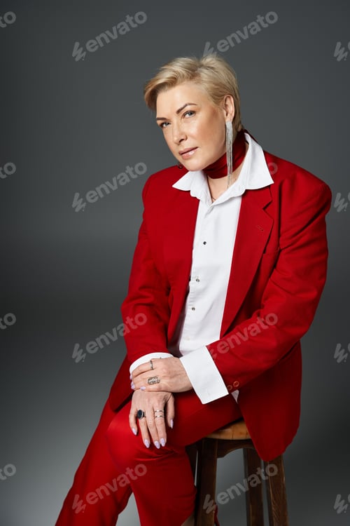 Preview: Elegant adult woman in stylish red attire poses confidently on a wooden stool indoors