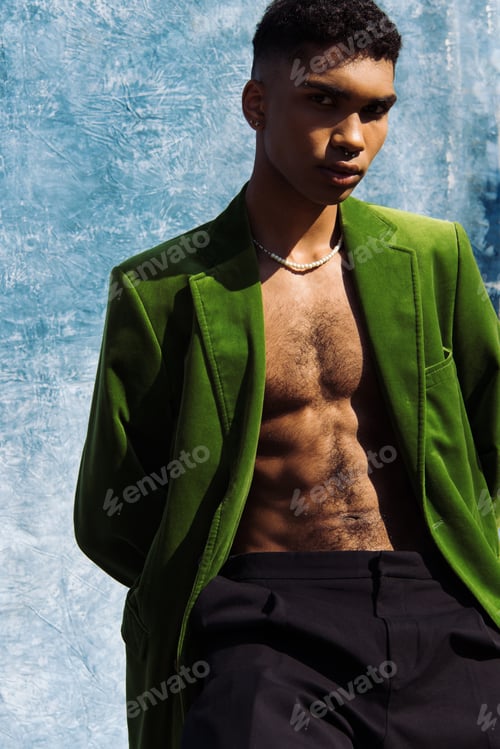 Preview: young african american man in green blazer and beads looking at camera near blue cloth on background