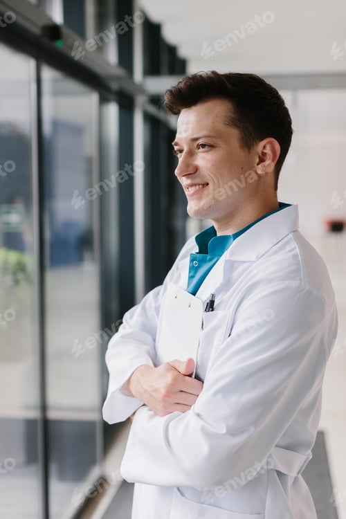 Preview: Male doctor with patient’s chart and pen posing near the window in a medical setting