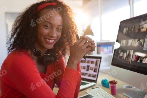 Preview: Remote worker wearing red sweater, headphones is holding pen on office desk: laptop, monitor, mug