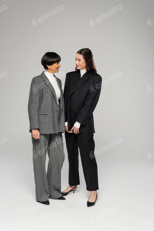 Preview: full length of cheerful asian mother and daughter in pantsuits looking at each other on grey
