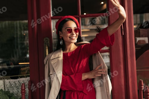 Preview: Happy tanned brunette woman in red dress, beret, trench coat and sunglasses smiles and waves hand i