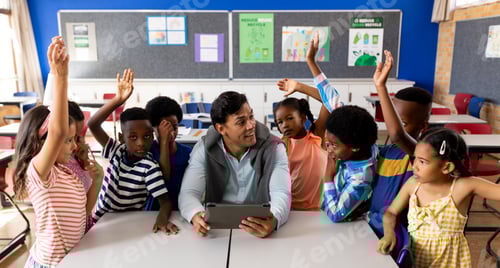 Preview: Diverse male teacher using tablet with children raising hands in class at elementary school