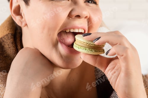 Preview: Cheerful woman is sitting in the kitchen and eating macarons cookies.
