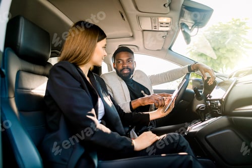 Preview: Business couple, pretty Caucasian woman and handsome African man, working in car with digital tablet