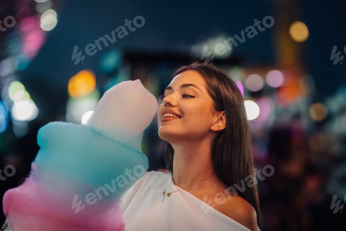 Preview: Happy young woman eating cotton candy at night in amusement park
