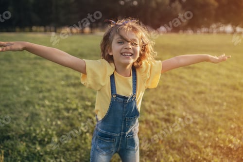 Preview: happy girl in yellow blouse denim overalls smiles spread arms as in flight on green grass in summer