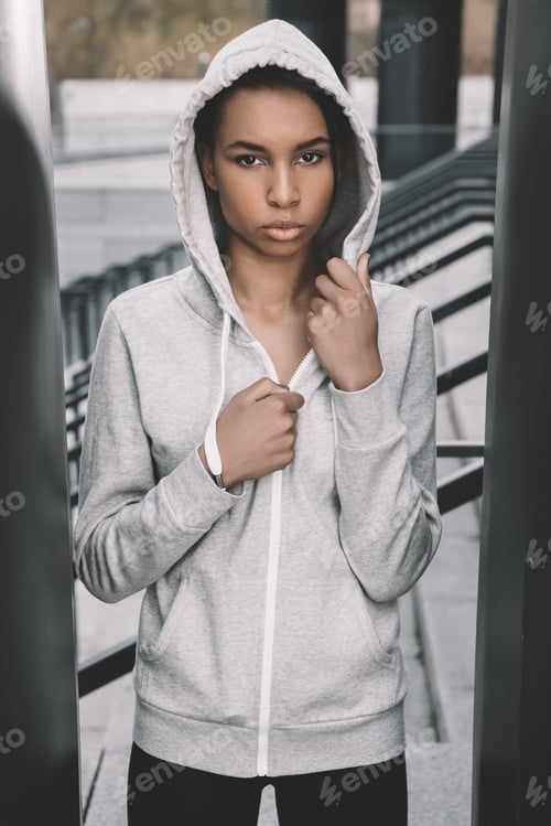 Preview: Attractive young fitness woman in sportswear wearing hood and posing on stadium