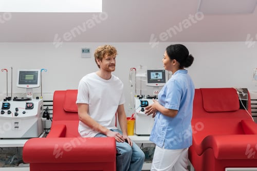 Preview: multiracial nurse in uniform talking to positive redhead volunteer sitting on medical chair