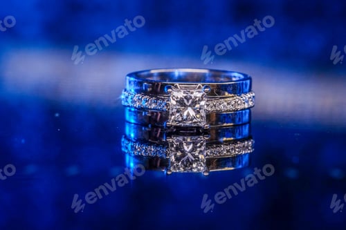 Preview: Closeup of a silver wedding ring with diamonds reflecting on a glass table under blue lights