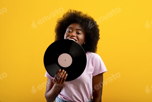 Preview: Joyful african american woman holding vinyl record, smiling and looking at camera on yellow