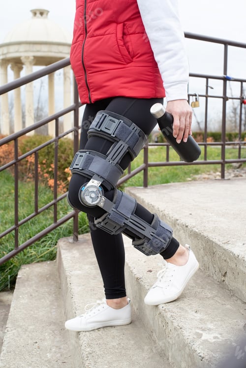Preview: Woman wearing knee brace or orthosis after leg surgery, walking in the park