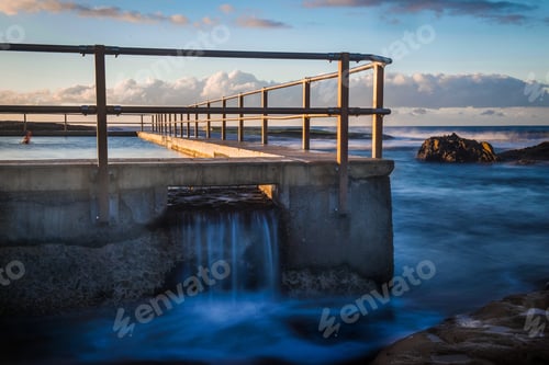Preview: Water flows through the overflow at a local rock pool. Sunrise lights up the clouds on the horizon