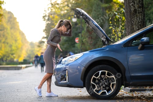 Preview: Puzzled female driver standing on a city street near her car with popped up hood looking at broken