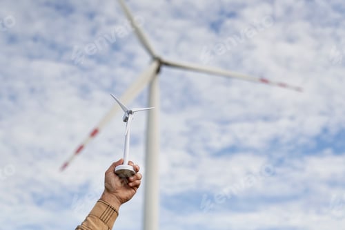 Preview: Hand of unrecognizable person holding a plastic windmill and real wind turbine in the background