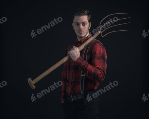 Preview: Young Fashionable Farmer In Red Checkered Shirt Holding Hayfork.