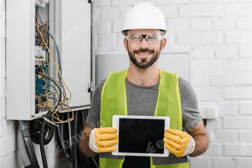 Preview: smiling handsome electrician showing tablet with blank screen near electrical box in corridor