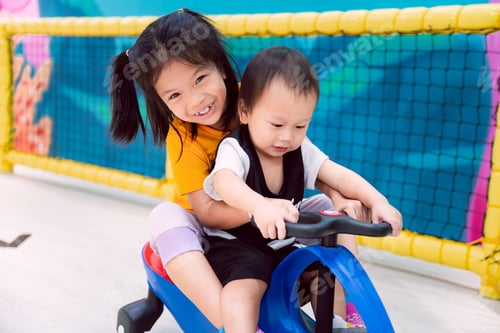 Preview: Asian boy and girl riding a blue toy car in the indoor playground with their family nearby.