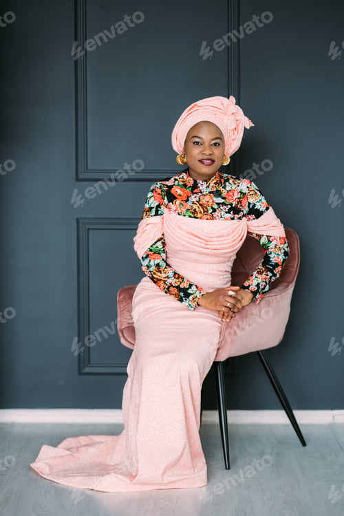 Preview: Pretty African woman wearing an pink fashionable traditional dress and headscarf, sitting on the