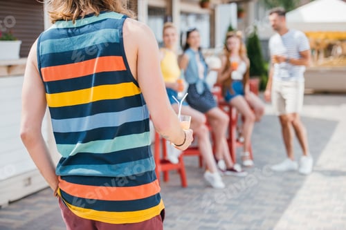 Preview: cropped shot of man holding glass with cocktail while friends sitting at beach bar