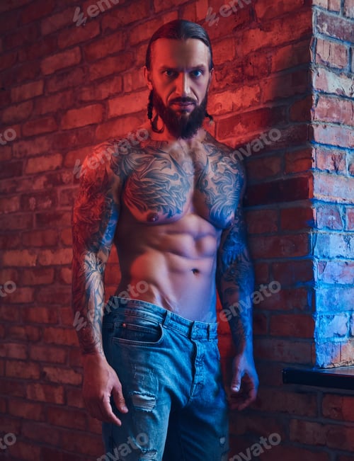 Preview: Handsome tattooed man with a stylish haircut and beard standing against a brick wall