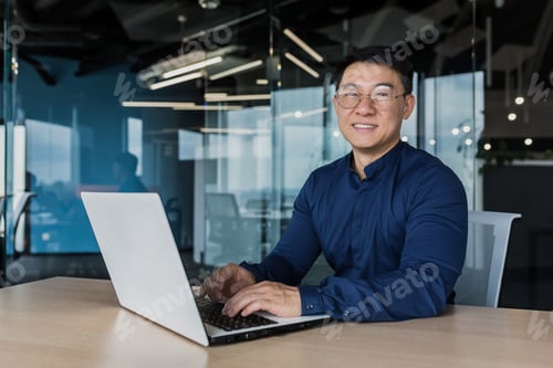 Preview: Young and successful asian businessman in glasses working on laptop inside modern office building