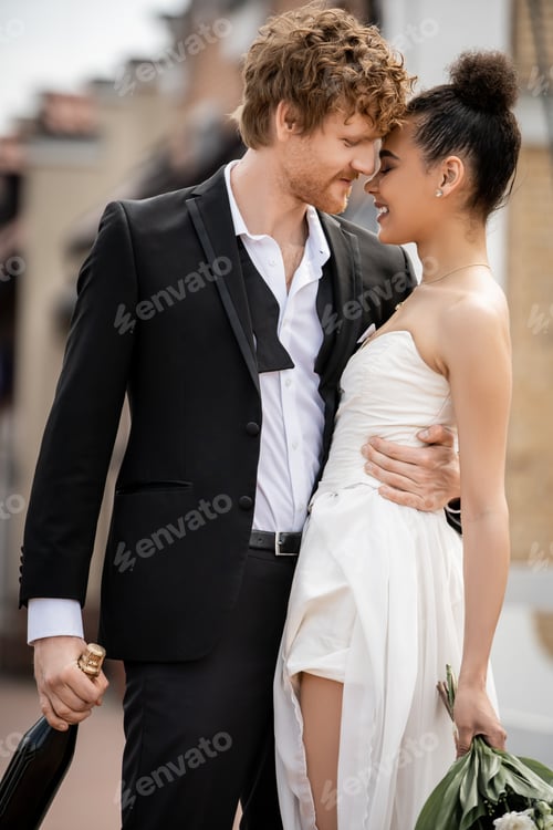 Preview: young redhead man in elegant suit embracing happy african american bride, wedding outdoors