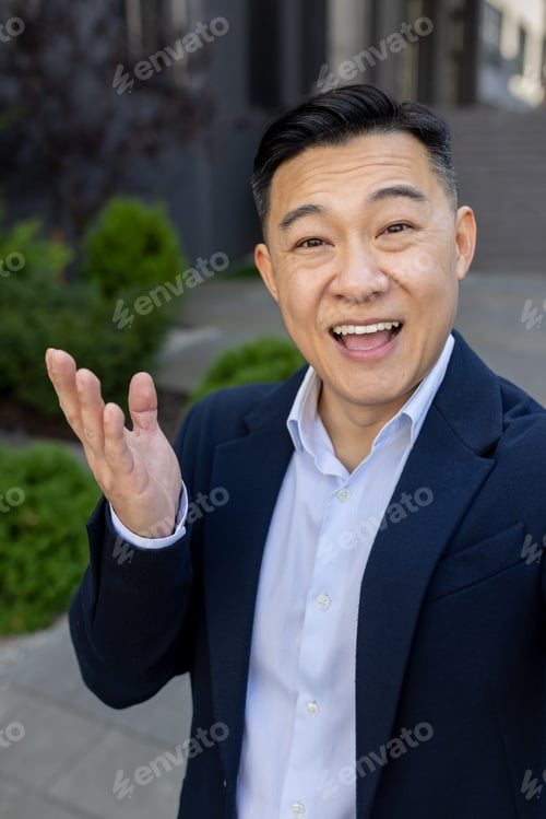 Preview: Happy Asian man standing outside in business suit and talking on video call gesturing with hands