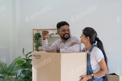 Preview: Portrait of happy couple indian in lounge of new home with key on move day surrounded by box