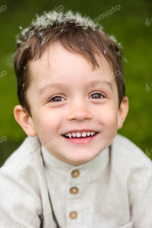 Preview: A cute boy in a linen shirt smiles with down from white dandelions on his head.