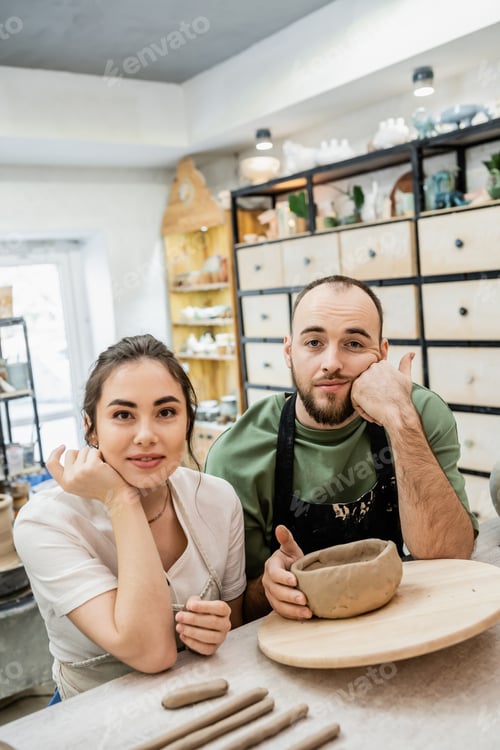 Preview: Smiling craftswoman in apron looking at camera near sad boyfriend and clay in ceramic workshop