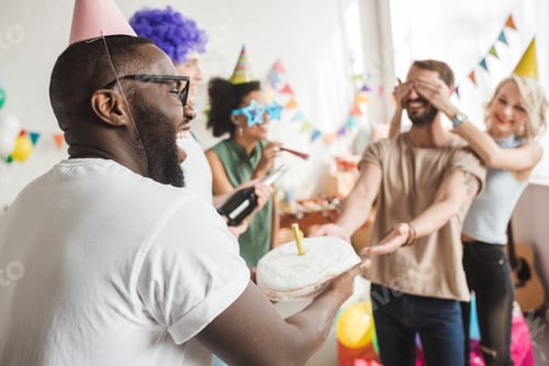Preview: Happy friends covering eyes of young man and greeting him with birthday cake