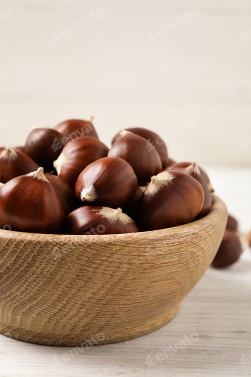 Preview: Fresh sweet edible chestnuts in bowl on white wooden table, closeup