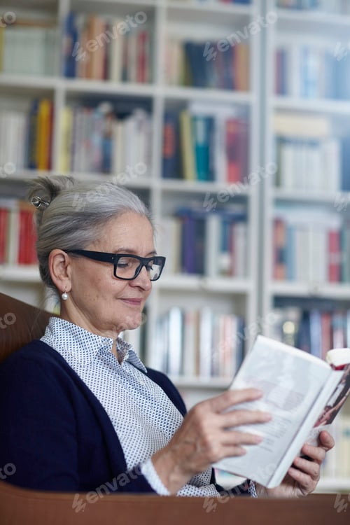 Preview: Grey haired mature woman reading book from bookshelves