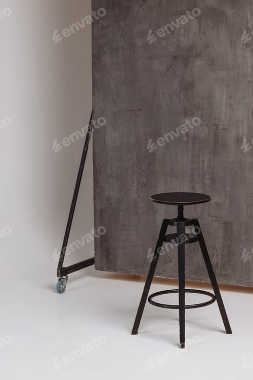 Preview: A black stool sits in front of a grey wall. Background photo studio with minimalist style.
