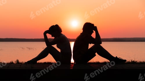 Preview: Silhouette of an upset couple sitting on the shore of the lake at sunset. Relationship problems