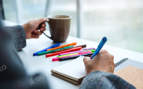 Preview: Closeup image of a woman writing on a blank notebook with colored pens while drinking coffee