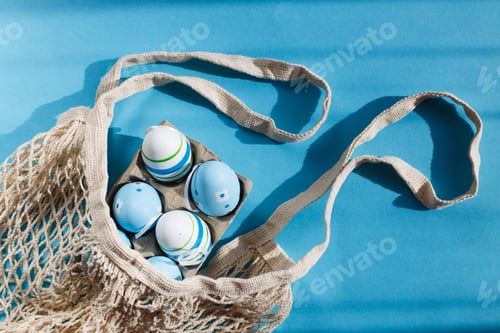 Preview: colorful handmade painted easter eggs in a string bag on blue background with hard shadows.