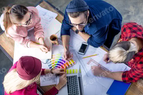 Preview: Overhead view of young male and female designers working at project with blueprints and devices